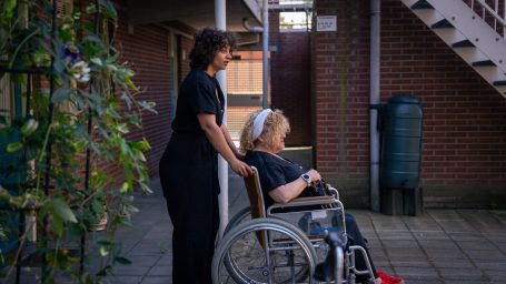 Vrouw in rolstoel met verzorgster buiten, naast een gevel onder een trap.