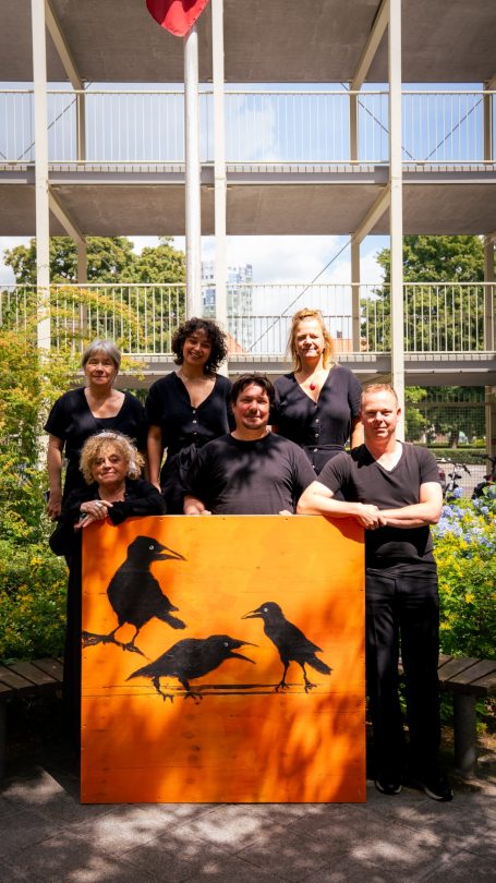 Zes mensen poseren voor een oranje bord met zwarte vogels in een groene omgeving.